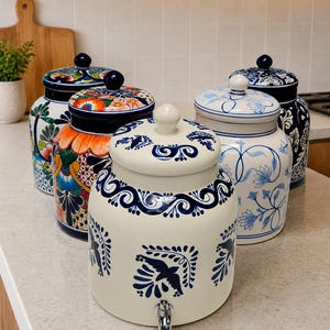 May include: A collection of five ceramic canisters with lids, including one cream-colored beverage dispenser with a silver spigot and blue bird patterns, alongside four decorative jars featuring various floral, botanical, and intricate blue and multi-colored designs.