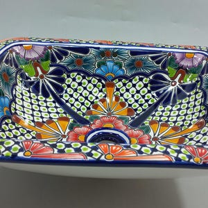 May include: Rectangular ceramic sink with a vibrant, hand-painted floral design. The sink features a white base and a colorful border with orange, blue, and green accents. The interior is adorned with intricate patterns.