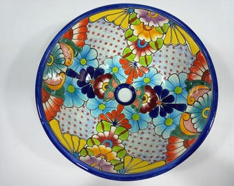 Op de afbeelding: Een handbeschilderde keramische wastafel met een levendig bloemmotief. De wastafel heeft een blauwe rand en een centrale afvoer. De bloemen zijn in tinten blauw, oranje, geel en paars, tegen een lichte achtergrond met kleine rode stippen.