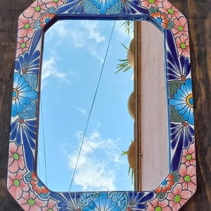 May include: An octagonal mirror with a floral border. The border features a repeating pattern of blue, pink, and green flowers on a white background. The mirror reflects a blue sky with clouds and a glimpse of a building.