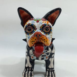 May include: A ceramic French bulldog figurine featuring a black base color with white skeletal bone patterns on the legs and chest. The face is painted with colorful floral and dot motifs around the eyes, a bright orange snout, and a red tongue.