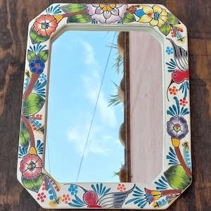 May include: A rectangular mirror with a white frame, adorned with colorful floral and hummingbird designs. The frame features red, orange, blue, and yellow flowers, with green leaves and a blue sky reflected in the mirror.