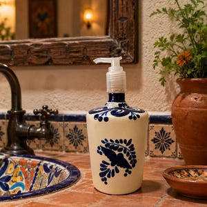 May include: A cream-colored ceramic soap dispenser featuring a hand-painted dark blue floral and leaf pattern. The cylindrical vessel is topped with a clear plastic pump mechanism designed for dispensing liquid soap or lotion in a bathroom setting.