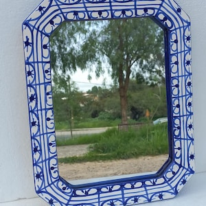 May include: An octagonal mirror with a white and blue decorative border. The border features a repeating pattern of blue lines and floral motifs. The mirror reflects a natural outdoor scene with trees and a dirt path.