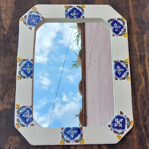 May include: Rectangular mirror with a cream-colored frame adorned with blue and yellow floral tile designs. The mirror reflects a blue sky with fluffy white clouds. The frame has an octagonal shape.