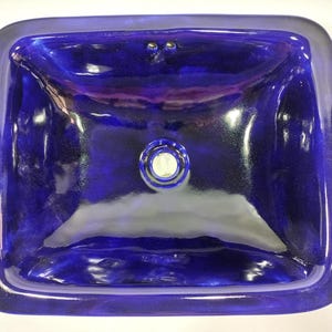 May include: A rectangular, deep blue glass sink with rounded corners. The sink has a glossy finish and a central drain with a silver and white fixture. Two small holes are visible near the top edge, likely for faucet installation.