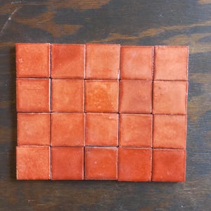 May include: A collection of twenty-four square, terracotta-colored tiles arranged in a rectangular grid. The tiles have a glossy finish and vary slightly in shade, creating a textured appearance. The tiles are set against a dark wood surface.