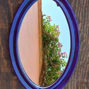 May include: An oval mirror with a deep blue frame. The mirror reflects a pink wall, a tree trunk, and a vibrant green tree with pink flowers against a blue sky. The mirror is mounted on a dark wood surface.