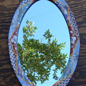 May include: Oval mirror with a blue, white, red, and yellow hand-painted ceramic frame. The frame features a floral design with intricate details. The mirror reflects a blue sky and a green tree.