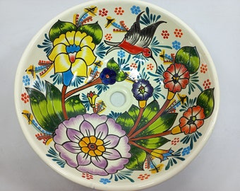 Op de afbeelding: Een witte keramische wastafel met een bloemen- en kolibrieontwerp. De wastafel heeft kleurrijke bloemen, groene bladeren en een rode kolibrie met zwarte vleugels. De wastafel is versierd met een witte achtergrond en een blauw, groen en rood kleurenschema.