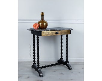 May include: A small, decorative table with a black base and ornate, turned legs. The tabletop is dark with a gold-painted drawer front. A gold-colored bottle and a pink flower sit on top. The table is approximately 30 inches long.