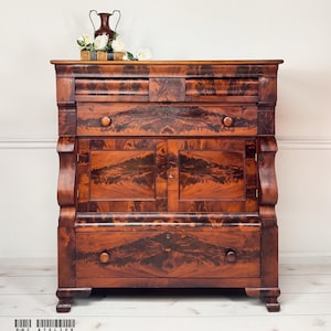 May include: A dark mahogany wood chest of drawers featuring a top drawer, a central cabinet with two doors, and a bottom drawer. The wood grain displays a symmetrical flame pattern. A small copper-colored vase and white flowers sit on top.