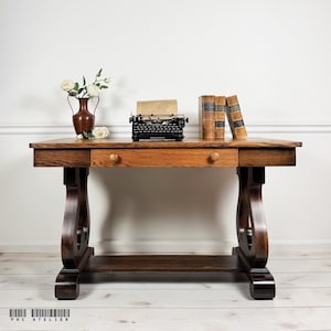 May include: A dark brown wooden desk with a single central drawer featuring two round knobs. The desk has curved side supports and a lower shelf. On top sits a black typewriter with paper, a brown vase with white flowers, and three vintage books.