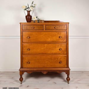 May include: A wooden chest of drawers with a warm brown finish and visible wood grain. The piece features two small top drawers and three full-width lower drawers, each with round wooden knobs. It stands on four turned legs with small casters.
