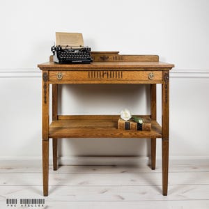 May include: A wooden writing desk with a single drawer featuring two brass pulls and decorative carvings on the legs. A black typewriter with a paper sheet sits on top, while a vintage book and a white flower rest on the lower shelf.