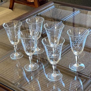 May include: Six clear crystal wine glasses with etched starburst patterns. The glasses have a classic design with a stemmed base and a flared bowl. They are arranged on a glass-topped table.