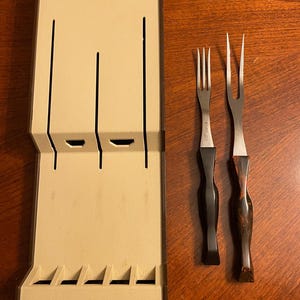 May include: A beige cutlery storage case with the word "Cutco" embossed on the front. Two serving forks with black and wood-grain handles are next to the case. The forks have stainless steel tines.