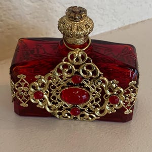 May include: A rectangular, ruby-red glass perfume bottle with a gold filigree design. The bottle features a crown-shaped stopper and is adorned with red jewels. The gold filigree wraps around the bottle, adding an ornate touch.