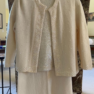 May include: A vintage cream-colored knit skirt suit. The set includes a collared jacket and a matching skirt. The jacket and the neckline of the top are embellished with sequins. The suit is displayed on a patterned chair.