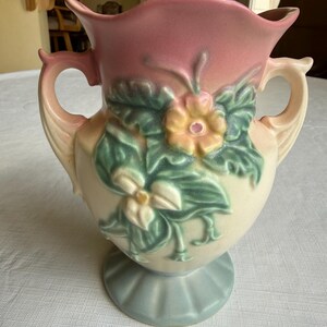 May include: A vintage ceramic vase with a pink top and cream-colored body. The vase features a floral design with green leaves and yellow flowers. It has two handles and a light blue base. A decorative piece for home decor.