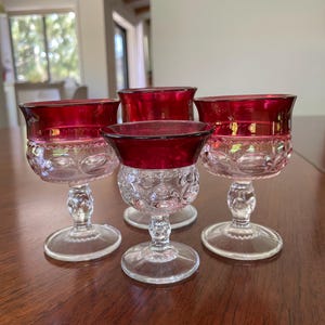 May include: Four vintage wine glasses with ruby red rims and clear, patterned bowls. The glasses have clear stems and bases, and are arranged on a dark wooden surface. The glasses are likely made of glass.