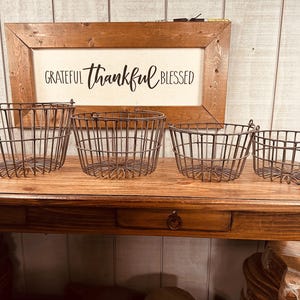 May include: Four metal wire baskets of varying sizes sit on a wooden table. The baskets are dark brown and have a rustic, farmhouse style. Behind the baskets is a framed sign with the words "Grateful, Thankful, Blessed" in black script.