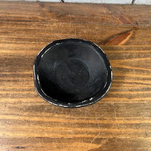 May include: A small, round, black ceramic bowl with an irregular, organic shape. The interior surface is matte black, while the outer rim features a distressed, white-painted finish that highlights the textured, uneven edge of the vessel.