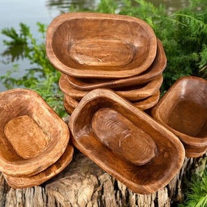 May include: A stack of hand-carved, wooden dough bowls in various sizes. The bowls have a rustic, natural wood grain finish and are arranged on a weathered wooden surface. The background features greenery and water, suggesting an outdoor setting.