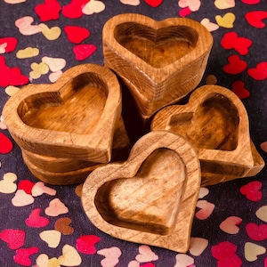 May include: Four wooden heart-shaped bowls stacked on top of each other. The bowls are made of natural wood and have a rustic finish. The bowls are arranged on a dark background with pink and white confetti.