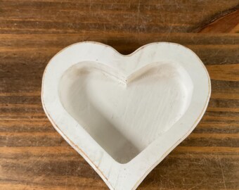 May include: A heart-shaped, white wooden bowl with a distressed finish. The bowl has a shallow interior and a slightly raised rim. The wood grain is visible, adding texture. The bowl is set against a wooden surface.