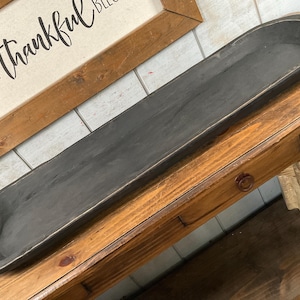 May include: A long, dark gray wooden dough bowl with rounded ends sits on a wooden table. The bowl has a distressed finish. A framed sign with the words "Grateful Thankful Blessed" is in the background.
