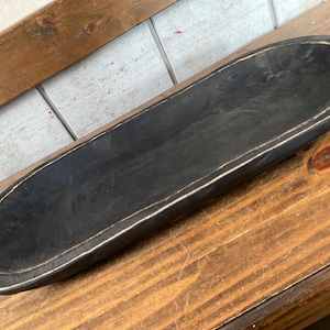 May include: A long, oval-shaped, black wooden bowl with a white trim. The bowl is set on a wooden surface. The interior of the bowl is black, and the rim has a distressed white finish. This decorative bowl is suitable for home decor.