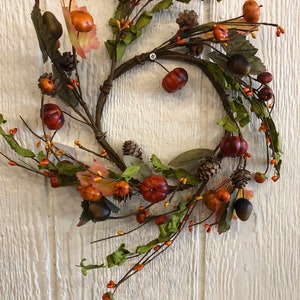 May include: A circular autumn wreath featuring miniature pumpkins, acorns, and pinecones. The wreath is made of brown twigs and adorned with green leaves and orange berries. The pumpkins are various shades of orange and red.