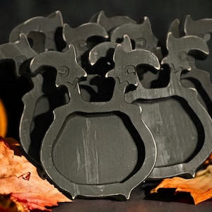 May include: A stack of dark gray metal coasters with a unique, stylized design. The coasters have a shield-like shape with a decorative top. A small orange pumpkin and autumn leaves are in the background, suggesting a fall theme.