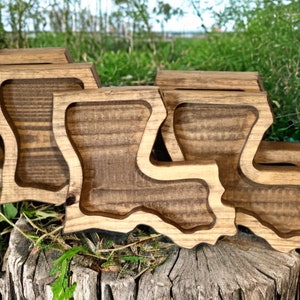 May include: A collection of wooden coasters, each carved in the shape of the state of Louisiana. The coasters are made of stained wood, showcasing the natural grain. They are arranged on a tree stump against a backdrop of green foliage.
