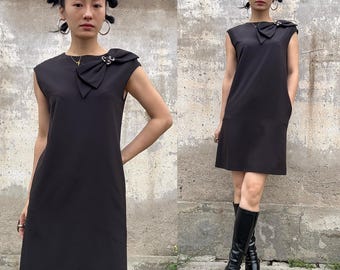 May include: A sleeveless black dress with a bow detail at the neckline. The dress has a simple, elegant design and falls to the mid-thigh. The bow is adorned with a silver-colored embellishment. The dress is paired with black knee-high boots.