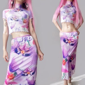 May include: A matching two-piece set comprising a crop top and a long skirt, both displaying a vivid purple and white floral pattern. The top has short sleeves and a high neckline, and the skirt has a flowing design. The outfit is crafted from a sheer, lightweight material.