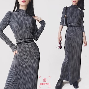 May include: A gray pleated dress with a high neckline and long sleeves. The dress features a black belt around the waist. The dress is floor-length and has a vertical striped pattern. The Instagram handle "BOOPVTG" is visible in the bottom right corner.