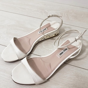 May include: A pair of white leather sandals with thin ankle straps. The sandals have a low heel with a clear, textured design. The brand name "miu miu" is visible on the insole.