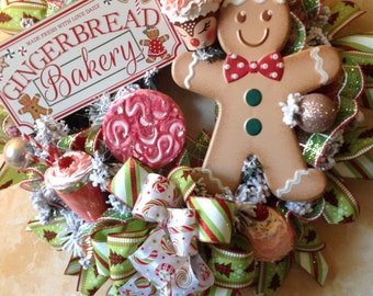Puede incluir: Una corona navideña decorada con cinta roja, verde y blanca, un hombre de jengibre, una galleta de jengibre, un cupcake y un letrero que dice "Gingerbread Bakery".