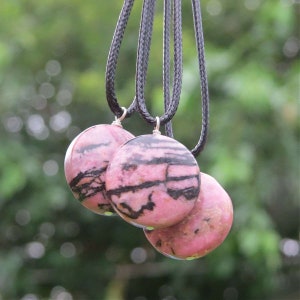 May include: Three pink and black patterned round pendants on black cords. The pendants are made of a natural stone with a pink and black marbled pattern.