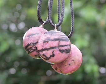 May include: Three pink and black patterned round pendants on black cords. The pendants are made of a natural stone with a pink and black marbled pattern.