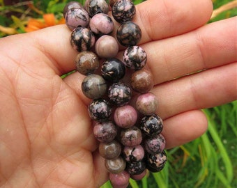 Puede incluir: Un brazalete de piedras preciosas rosa, negro y marrón con cuentas redondas. El brazalete está hecho de piedras naturales y tiene una longitud aproximada de 20 cm.