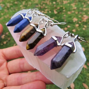 May include: Three pairs of silver crystal earrings with different colored stones. The earrings feature a silver teardrop-shaped setting with a pointed crystal stone. The stones are blue, brown, and purple.