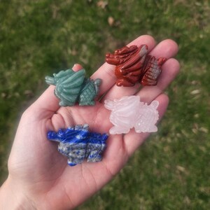 May include: Four carved stone dragon figurines in green, red, blue, and pink hues. The dragons are detailed with wings and textured bodies. The figurines are held in a hand against a blurred green background.