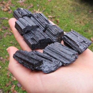 Puede incluir: Una colecci&oacute;n de cristales de turmalina negra en bruto. Los cristales, sostenidos en una mano, muestran superficies rugosas y texturizadas y un color oscuro natural. Las piezas de turmalina var&iacute;an en tama&ntilde;o y forma, con estr&iacute;as visibles.