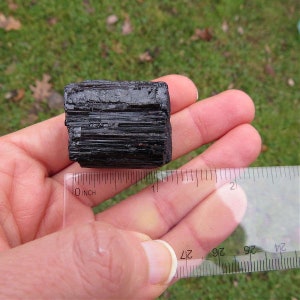 Puede incluir: Un cristal de turmalina negra en bruto sostenido en una mano, con una regla que muestra su tama&ntilde;o. El cristal es de color oscuro y opaco con estr&iacute;as visibles. La regla indica aproximadamente 2,5 cm de longitud.