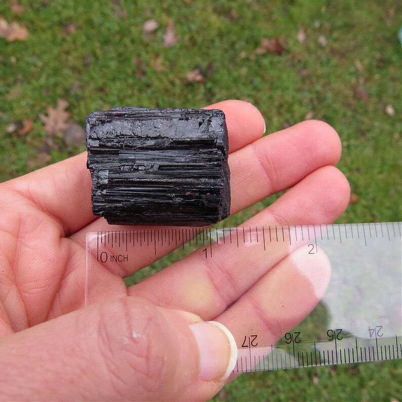 Puede incluir: Un cristal de turmalina negra en bruto sostenido en una mano, con una regla que muestra su tama&ntilde;o. El cristal es de color oscuro y opaco con estr&iacute;as visibles. La regla indica aproximadamente 2,5 cm de longitud.