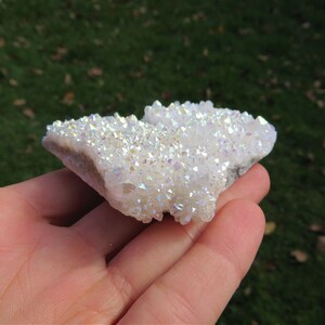 Puede incluir: Un c&uacute;mulo de cristales de cuarzo blanco iridiscente con una textura natural y rugosa. Los cristales est&aacute;n agrupados en una forma plana e irregular, y tienen una apariencia brillante, como un arco&iacute;ris.