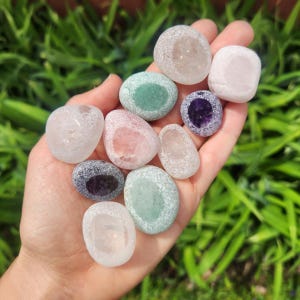 May include: A hand holds a collection of polished gemstones in various colors, including pink, green, purple, and clear. The stones have a smooth, rounded shape and are displayed against a backdrop of green foliage.